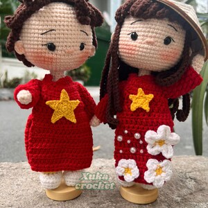 May include: Two handmade crochet dolls, one male and one female, holding hands. The dolls wear red dresses with yellow star accents. The female doll has a conical hat and white flower details. The dolls are on wooden stands. The text "Xuka crochet" is visible.