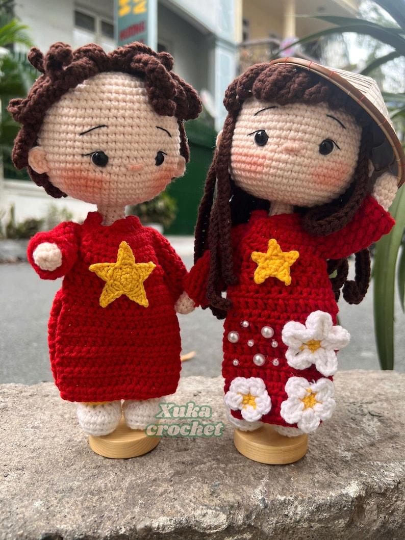 May include: Two handmade crochet dolls, one male and one female, holding hands. The dolls wear red dresses with yellow star accents. The female doll has a conical hat and white flower details. The dolls are on wooden stands. The text "Xuka crochet" is visible.