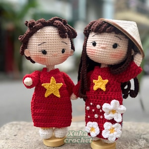 May include: Two handmade crochet dolls, a boy and a girl, holding hands. They wear red dresses with yellow star accents. The girl doll has a conical hat and white flower details. The text "Xuka crochet" is visible.
