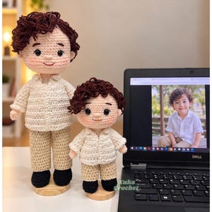 May include: Two handmade crochet dolls with dark brown curly hair, wearing cream-colored button-down shirts and tan trousers with black shoes. The dolls are positioned next to a laptop displaying a photograph of a child wearing the same outfit.