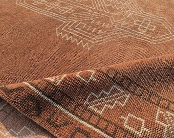 Puede incluir: Alfombra de color terracota con un diseño geométrico blanco. La textura de la alfombra es visible, y una parte está doblada, revelando la parte inferior con un patrón similar. El diseño presenta formas y líneas angulares.