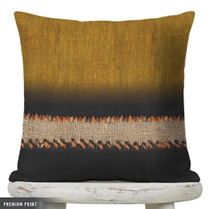 May include: Square decorative pillow with a textured design. The top is a golden-yellow color, transitioning to black at the bottom. A band of burlap-like material with orange stitching accents the design. The pillow is on a white stool.