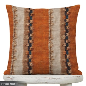 May include: Square decorative pillow with a textured orange, beige, and brown striped pattern. The stripes are vertical, with a woven texture and dark blue accents. The pillow is on a white wooden surface.