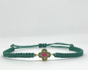 May include: A teal cord bracelet featuring a central gold-toned clover-shaped charm. The charm is inlaid with a bright pink center stone surrounded by small teal crystals. The bracelet has an adjustable macrame closure and the text Ostei.
