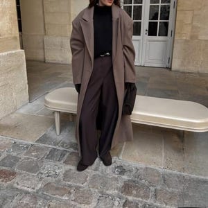 May include: A long, light brown coat paired with dark brown trousers and a black turtleneck. The outfit is accessorized with a black belt, gloves, and a small handbag. The ensemble is completed with dark brown shoes.