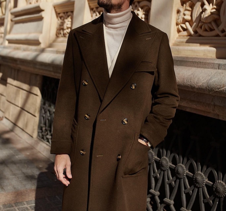 May include: A dark brown double-breasted wool coat with wide lapels and six buttons, worn over a cream-colored ribbed turtleneck sweater. The coat features long sleeves, two side pockets, and a structured fit with a classic tailored silhouette.