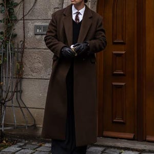 Könnte beinhalten: Ein langer, dunkelbrauner Mantel mit klassischem Design. Der Mantel hat ein fallendes Revers und einen Knopfverschluss. Die Person trägt einen schwarzen Pullover, ein weißes Hemd und eine gemusterte Krawatte. Schwarze Handschuhe werden getragen.