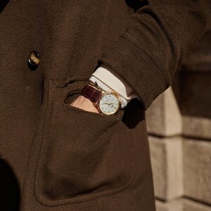 May include: A gold-tone wristwatch with a white dial and a dark brown leather strap featuring a crocodile-embossed texture. The watch is worn on a wrist partially tucked into the pocket of a dark brown wool coat over a white ribbed sweater.