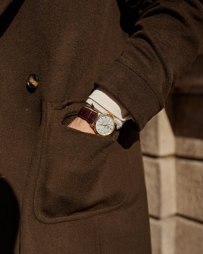 May include: A gold-tone wristwatch with a white dial and a dark brown leather strap featuring a crocodile-embossed texture. The watch is worn on a wrist partially tucked into the pocket of a dark brown wool coat over a white ribbed sweater.