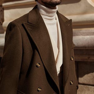 May include: A dark olive green double-breasted coat with wide lapels and brown buttons, worn over a cream-colored ribbed turtleneck sweater and a light-colored collared shirt underneath, showcasing a layered texture and structured tailoring.