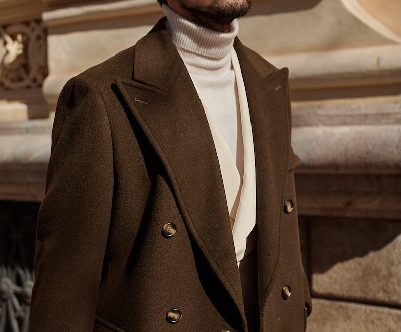 May include: A dark olive green double-breasted coat with wide lapels and brown buttons, worn over a cream-colored ribbed turtleneck sweater and a light-colored collared shirt underneath, showcasing a layered texture and structured tailoring.