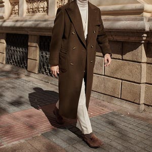 May include: A long, double-breasted dark brown wool coat with wide lapels and brown buttons, worn over a cream-colored turtleneck sweater, light-colored trousers, and brown suede loafers. The coat features two large patch pockets on the front.