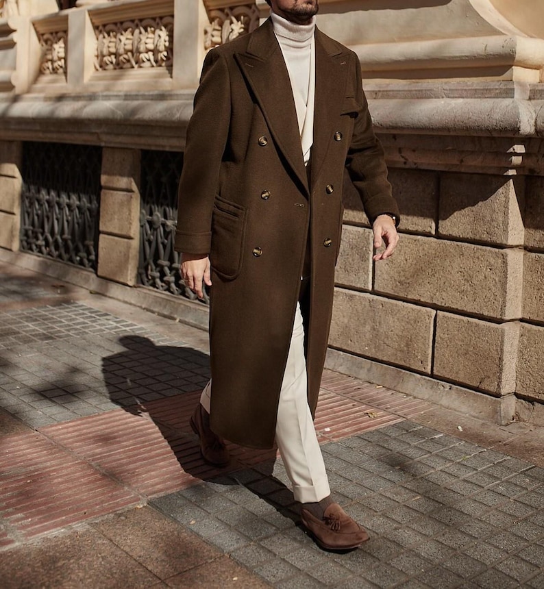 May include: A long, double-breasted dark brown wool coat with wide lapels and brown buttons, worn over a cream-colored turtleneck sweater, light-colored trousers, and brown suede loafers. The coat features two large patch pockets on the front.