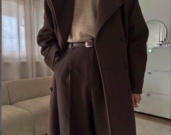 May include: A dark brown double-breasted wool coat worn over a beige crew-neck sweater and dark brown trousers. A thin brown leather belt with a silver-toned buckle is fastened at the waist. The coat features wide lapels and dark buttons.