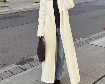 May include: A long, cream-colored coat with a double-breasted design. The coat is worn over a black sweater and blue jeans. A dark brown tote bag is held in the left hand. The person is wearing black shoes.