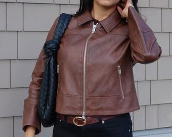 May include: A brown leather jacket with a collared neckline and a silver zipper. The jacket has two zippered pockets and is paired with a black woven shoulder bag and a brown belt with a gold buckle.