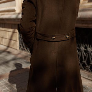 May include: A dark brown long coat featuring a back waist tab with two dark buttons. The coat is constructed from a smooth, solid-colored fabric and includes vertical seam detailing along the back panel, extending from the waist to the hem.