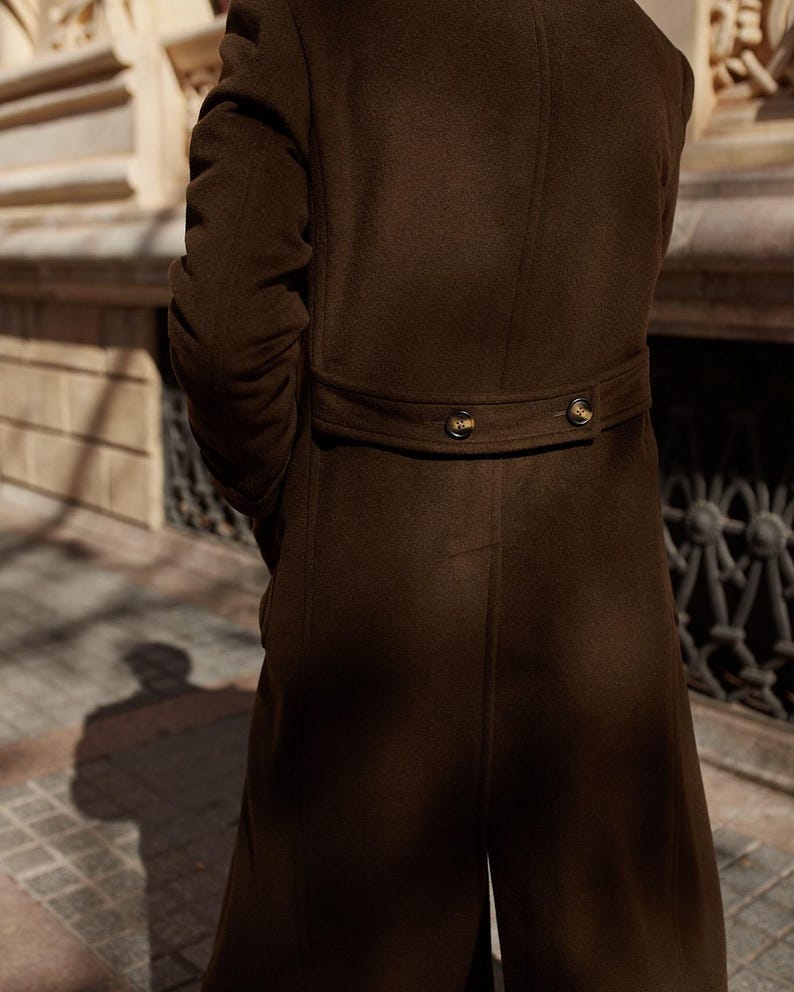 May include: A dark brown long coat featuring a back waist tab with two dark buttons. The coat is constructed from a smooth, solid-colored fabric and includes vertical seam detailing along the back panel, extending from the waist to the hem.