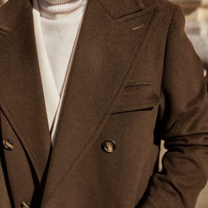 May include: A dark brown double-breasted coat with wide lapels and dark buttons, worn over a cream-colored turtleneck sweater. The coat features a structured collar, a chest pocket, and a textured fabric finish, providing a layered appearance.