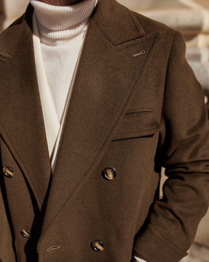 May include: A dark brown double-breasted coat with wide lapels and dark buttons, worn over a cream-colored turtleneck sweater. The coat features a structured collar, a chest pocket, and a textured fabric finish, providing a layered appearance.