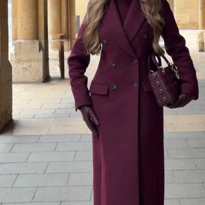 Peut inclure: Un long manteau bordeaux à double boutonnage, avec un sac à main et des gants assortis. Le manteau présente un revers classique, deux rangées de boutons et une coupe ajustée. Le sac à main a un design matelassé.