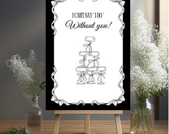 Könnte beinhalten: Ein schwarz-weißes Schild auf einer hölzernen Staffelei mit dem Text "I Can't Say 'I Do' Without you!" und einer Zeichnung von Sektgläsern. Das Schild ist mit einem weißen Schleifendesign umrahmt. Weiße Blumen befinden sich in Glasvasen auf beiden Seiten.