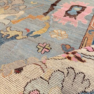 May include: A woven area rug featuring a light blue background with a multi-colored floral and geometric pattern in shades of pink, orange, cream, and dark brown. The rug is folded over to reveal a cream-colored underside with a textured weave.