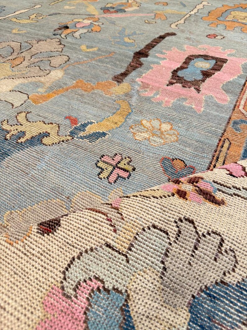 May include: A woven area rug featuring a light blue background with a multi-colored floral and geometric pattern in shades of pink, orange, cream, and dark brown. The rug is folded over to reveal a cream-colored underside with a textured weave.