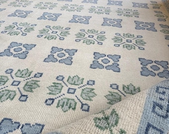 May include: A rectangular area rug featuring an off-white base with a repeating grid pattern of blue and green floral motifs. The rug has a wide blue border with a geometric pattern and a fringe edge. One corner is folded over to show the texture.