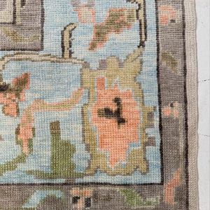 May include: A woven area rug featuring a light blue background with a repeating floral pattern in shades of peach, olive green, and dark gray. The rug has a wide, dark gray border with small geometric accents in peach and light blue throughout.