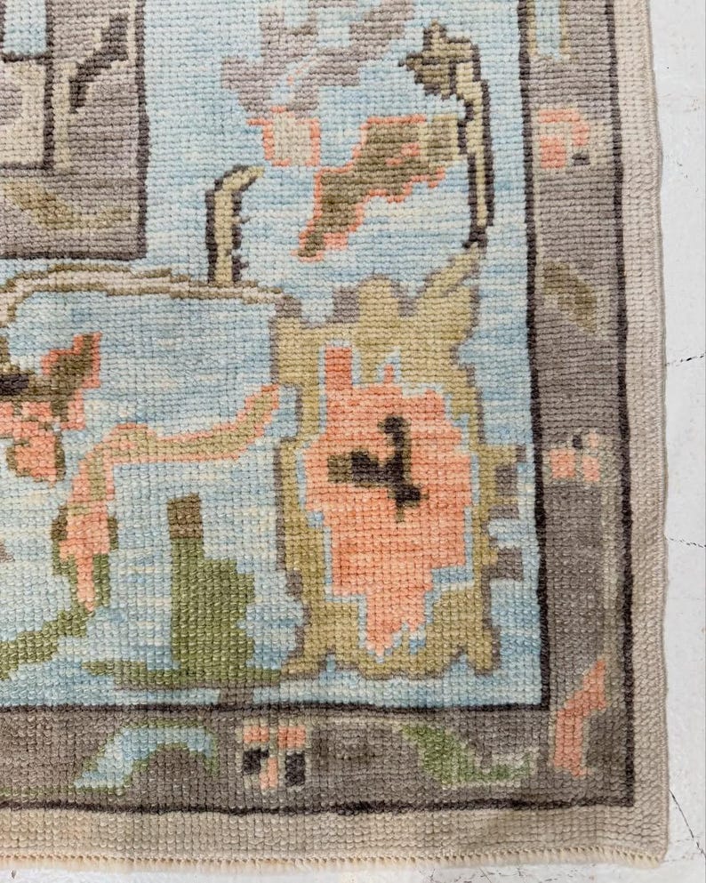 May include: A woven area rug featuring a light blue background with a repeating floral pattern in shades of peach, olive green, and dark gray. The rug has a wide, dark gray border with small geometric accents in peach and light blue throughout.