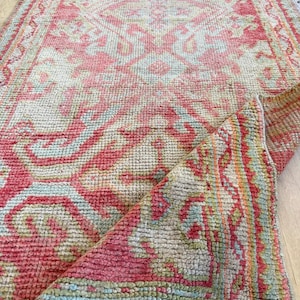 May include: A rectangular area rug featuring a woven pattern in shades of coral pink, light blue, and cream. The rug has a textured pile surface with a folded corner revealing the underside, displaying a repeating geometric and floral motif.