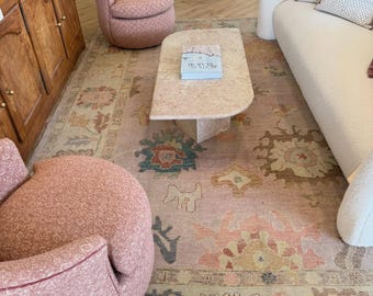 May include: A large rectangular area rug featuring a faded floral and geometric pattern in muted tones of pink, tan, blue, and brown. The rug is positioned under a light-colored curved sofa, two rounded pink textured chairs, and a stone coffee table.