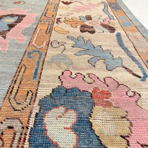 May include: A hand-knotted area rug featuring a light blue central field and a wide cream-colored border. The design includes intricate floral and leaf motifs in shades of pink, orange, dark blue, and tan, with a geometric orange border trim.