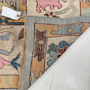 May include: A woven area rug with a cream background featuring a repeating pattern of stylized pink animals, blue floral motifs, and orange geometric borders. The rug is folded back to reveal a light blue edge and a white paper tag with text.