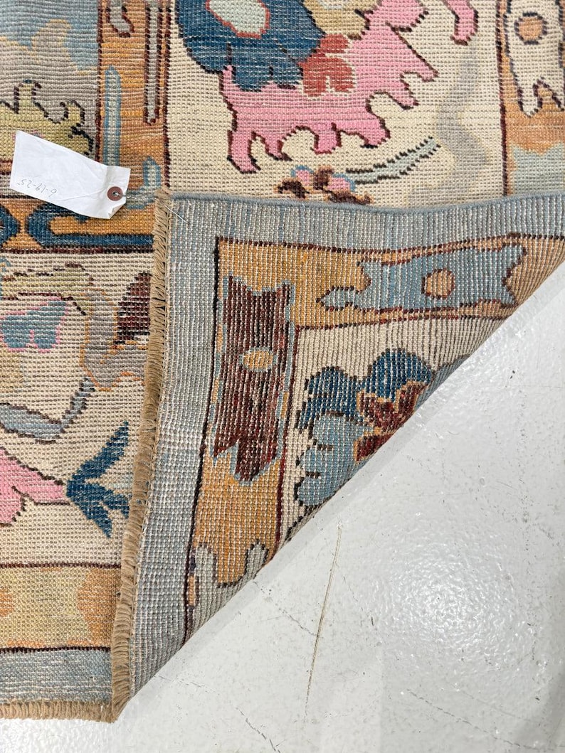 May include: A woven area rug with a cream background featuring a repeating pattern of stylized pink animals, blue floral motifs, and orange geometric borders. The rug is folded back to reveal a light blue edge and a white paper tag with text.