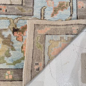 May include: A woven area rug with a light blue background featuring a floral pattern in shades of peach, olive green, and brown. The rug is folded at the corner to reveal the underside, showing a light beige woven texture and a stitched edge finish.