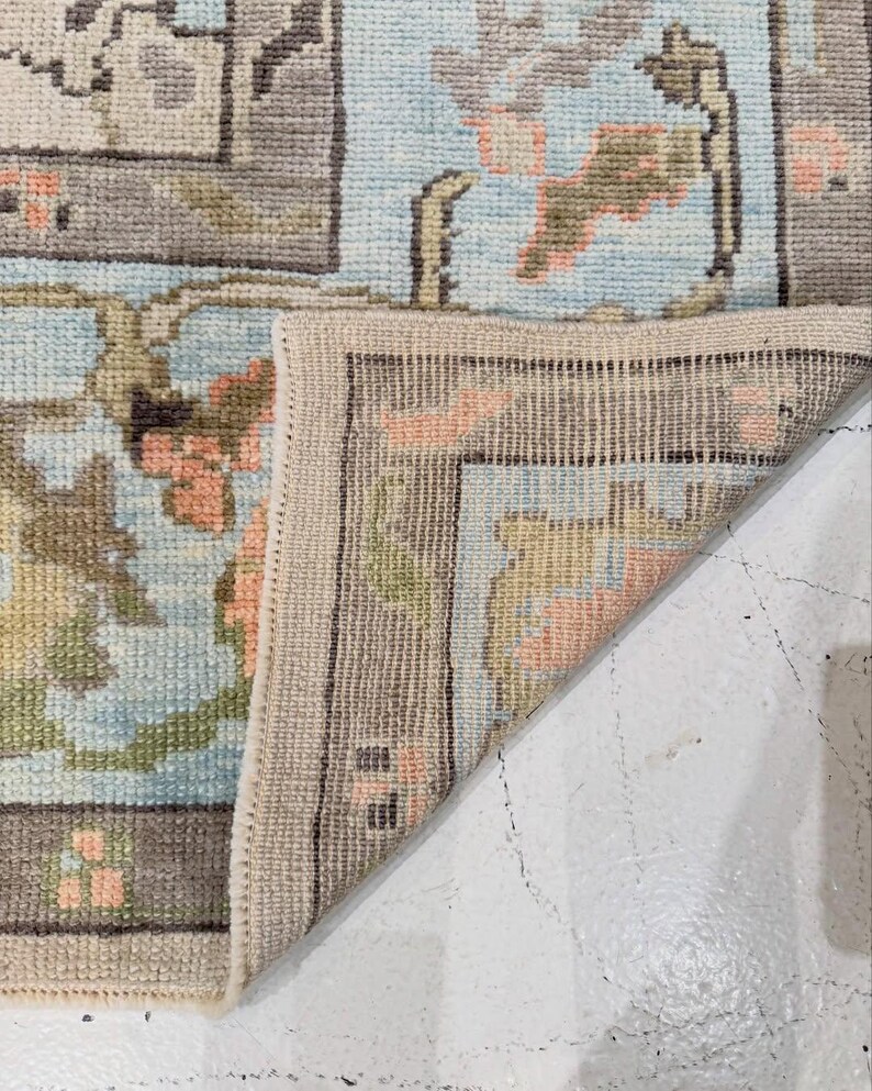 May include: A woven area rug with a light blue background featuring a floral pattern in shades of peach, olive green, and brown. The rug is folded at the corner to reveal the underside, showing a light beige woven texture and a stitched edge finish.