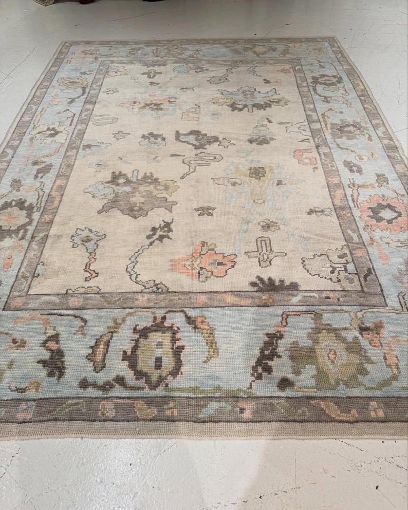 May include: A rectangular area rug featuring a cream-colored field with a central medallion and scattered floral motifs in shades of light blue, muted brown, and soft peach. The rug is framed by a wide border with repeating floral patterns.