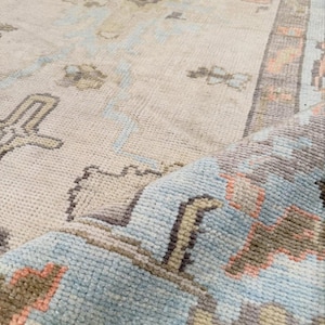 May include: A woven area rug featuring a cream-colored base with a repeating pattern in shades of light blue, soft coral, and muted brown. The rug has a visible pile texture and a folded corner that reveals the underside and the intricate weave.