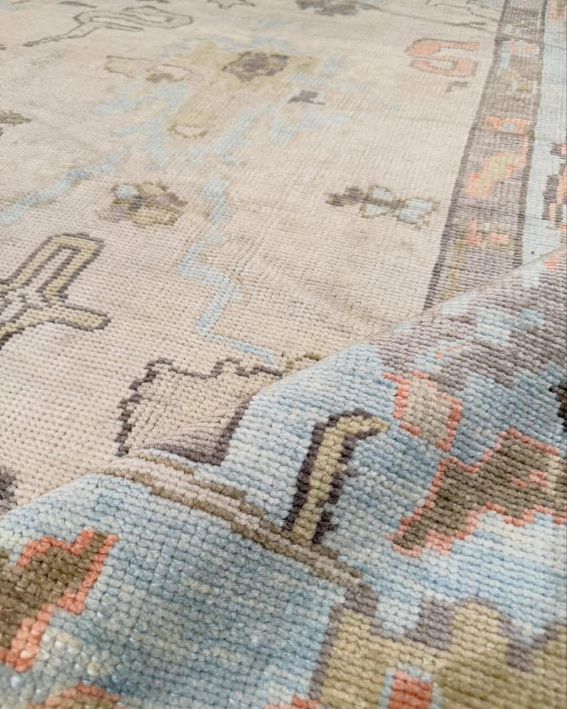May include: A woven area rug featuring a cream-colored base with a repeating pattern in shades of light blue, soft coral, and muted brown. The rug has a visible pile texture and a folded corner that reveals the underside and the intricate weave.