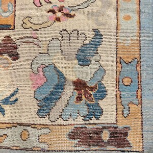 May include: A woven area rug featuring a cream-colored field with a central blue and gray floral motif, surrounded by a border of orange, blue, and brown geometric shapes. The rug has a light blue outer edge and a short, tan-colored fringe at the bottom.