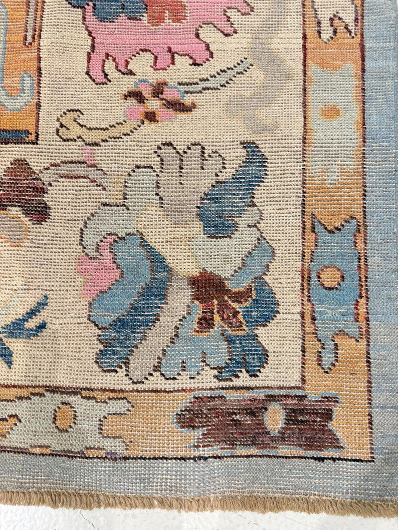May include: A woven area rug featuring a cream-colored field with a central blue and gray floral motif, surrounded by a border of orange, blue, and brown geometric shapes. The rug has a light blue outer edge and a short, tan-colored fringe at the bottom.