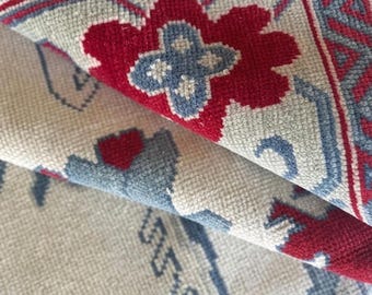 May include: A woven rug featuring a cream-colored base with a repeating pattern of red floral motifs and light blue geometric borders. The rug is folded over to reveal the underside, showing the dense, textured weave of the fabric construction.