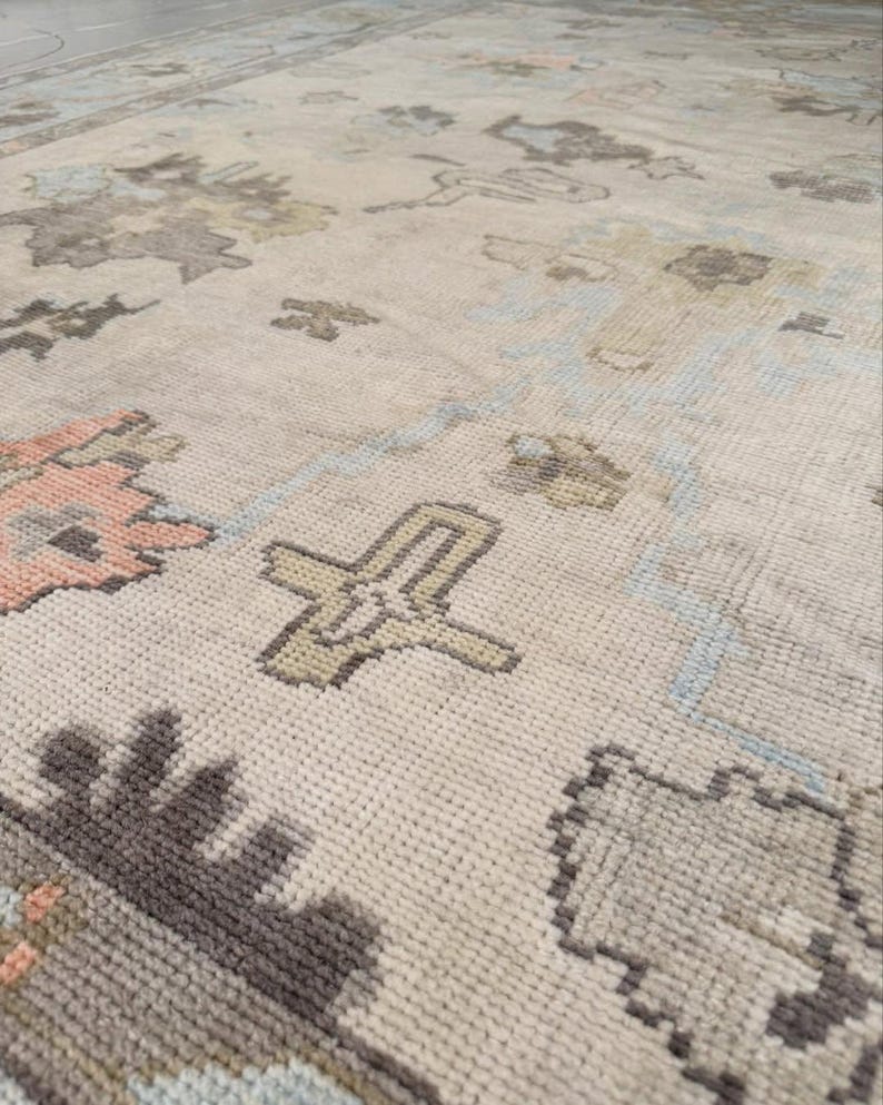 May include: A woven area rug featuring a light beige base with a repeating pattern of stylized floral and geometric motifs in muted shades of charcoal gray, soft salmon pink, pale blue, and olive green across the textured surface.