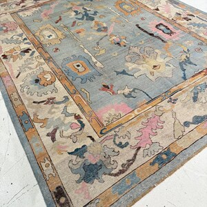 May include: A rectangular area rug featuring a light blue central field with a multi-colored floral and geometric pattern. The border displays a repeating design in shades of tan, pink, blue, and cream, framing the intricate central motifs.