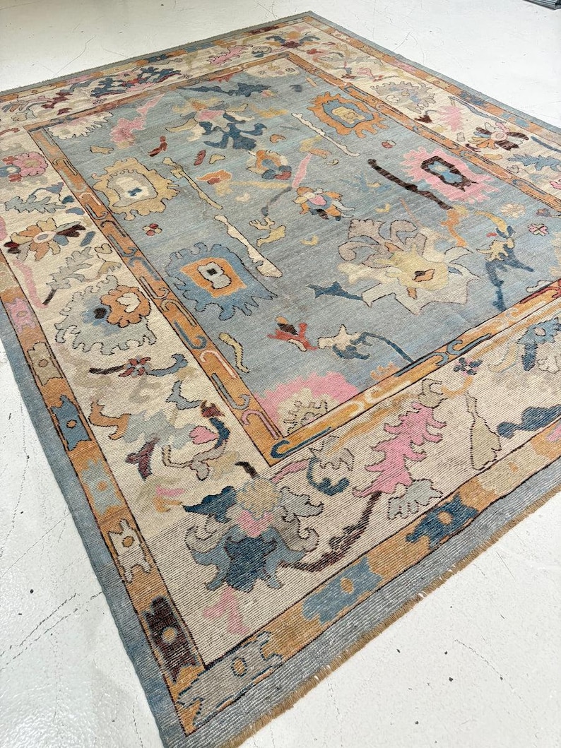 May include: A rectangular area rug featuring a light blue central field with a multi-colored floral and geometric pattern. The border displays a repeating design in shades of tan, pink, blue, and cream, framing the intricate central motifs.