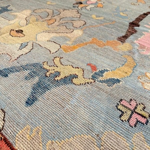May include: A light blue area rug featuring a repeating pattern of stylized floral and abstract motifs in shades of yellow, pink, dark blue, and brown. The woven textile displays a textured surface with visible knots and intricate, colorful designs.