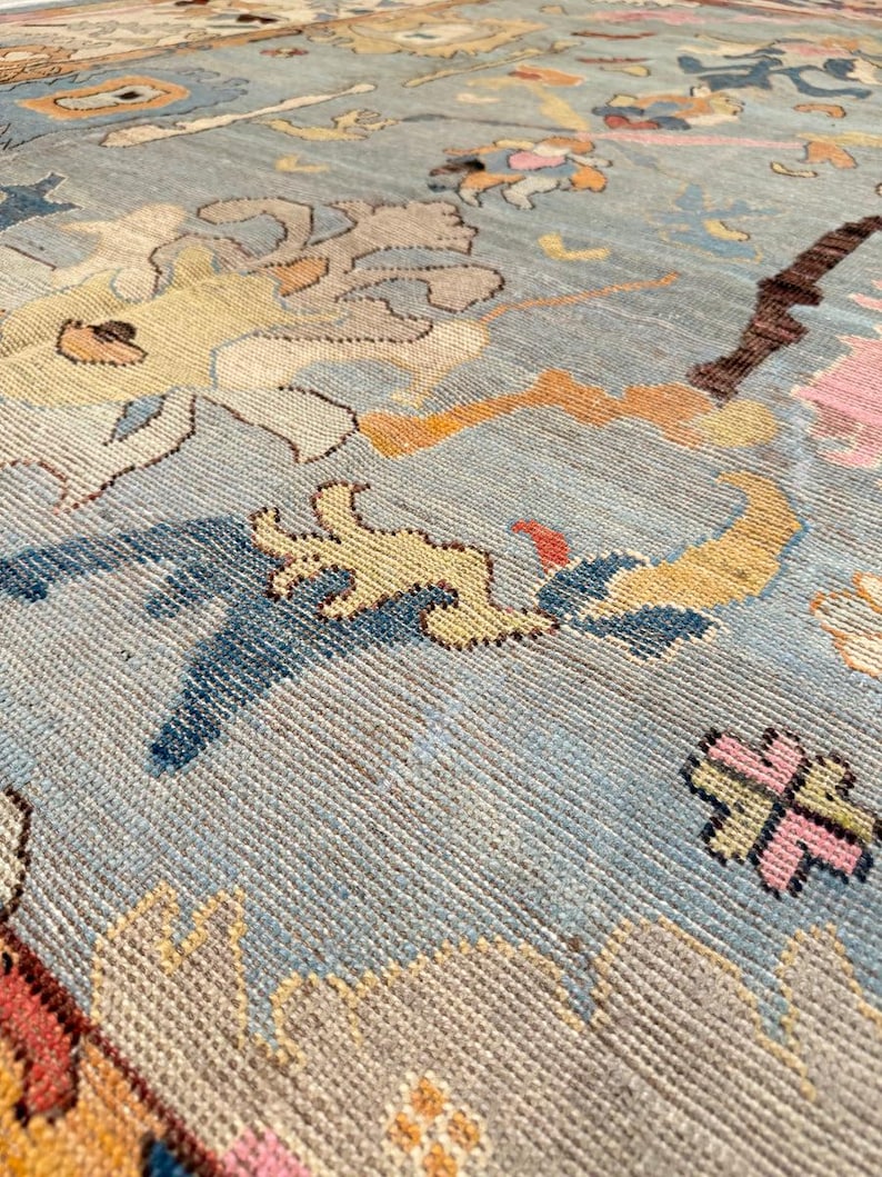 May include: A light blue area rug featuring a repeating pattern of stylized floral and abstract motifs in shades of yellow, pink, dark blue, and brown. The woven textile displays a textured surface with visible knots and intricate, colorful designs.
