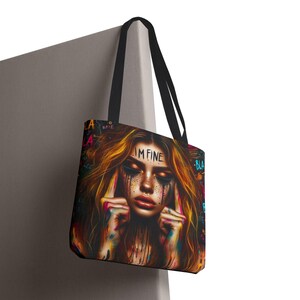 Op de afbeelding: Een zwarte tote bag met een kleurrijke grafische print van een persoon met lang haar, gesloten ogen en verfachtige strepen op het gezicht. De tekst "I'M FINE" staat op het voorhoofd, omringd door de tekst "BLA" in verschillende kleuren.